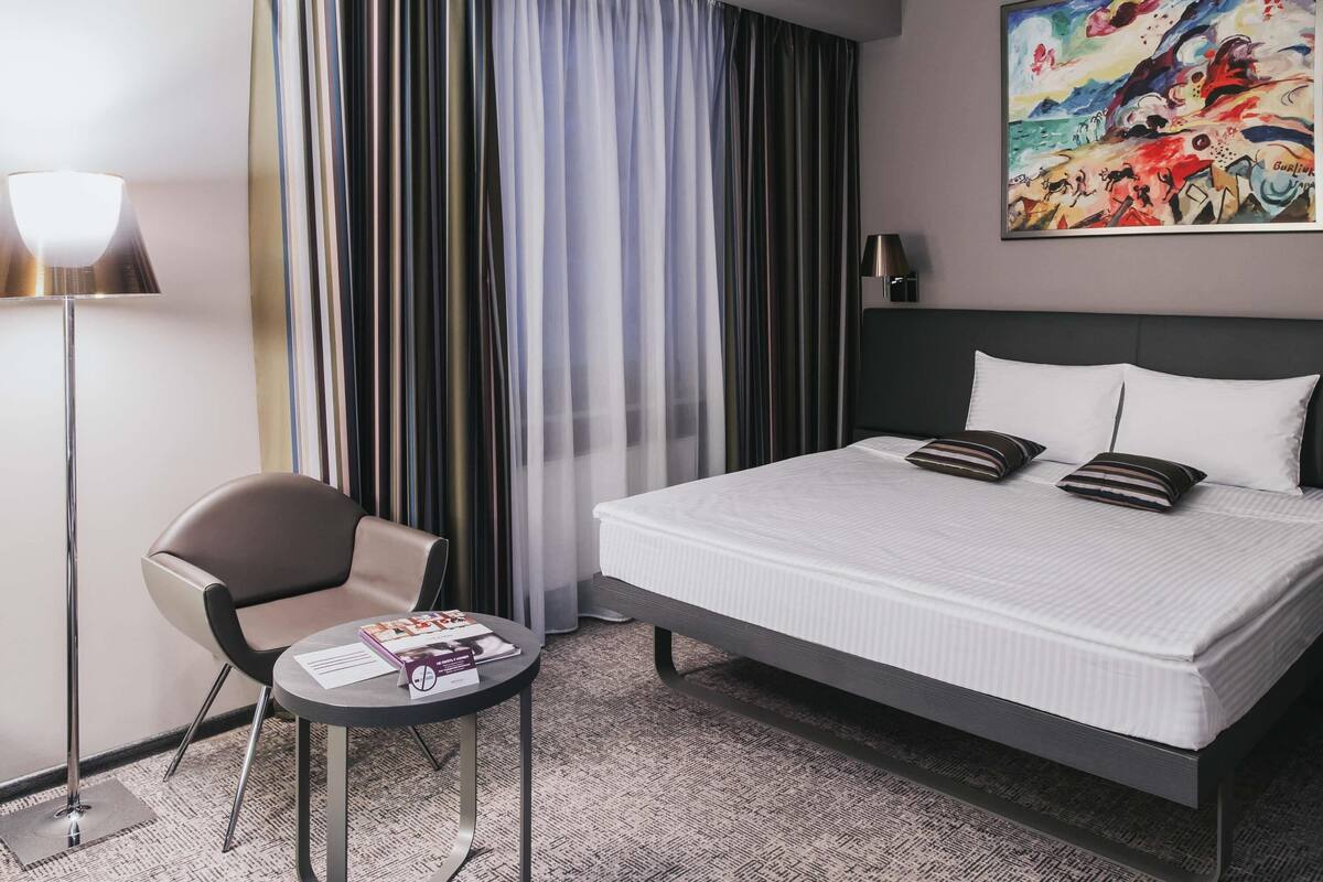 Ett hotellrum med en säng, en stol, ett litet bord, en lampa och en tavla på väggen.