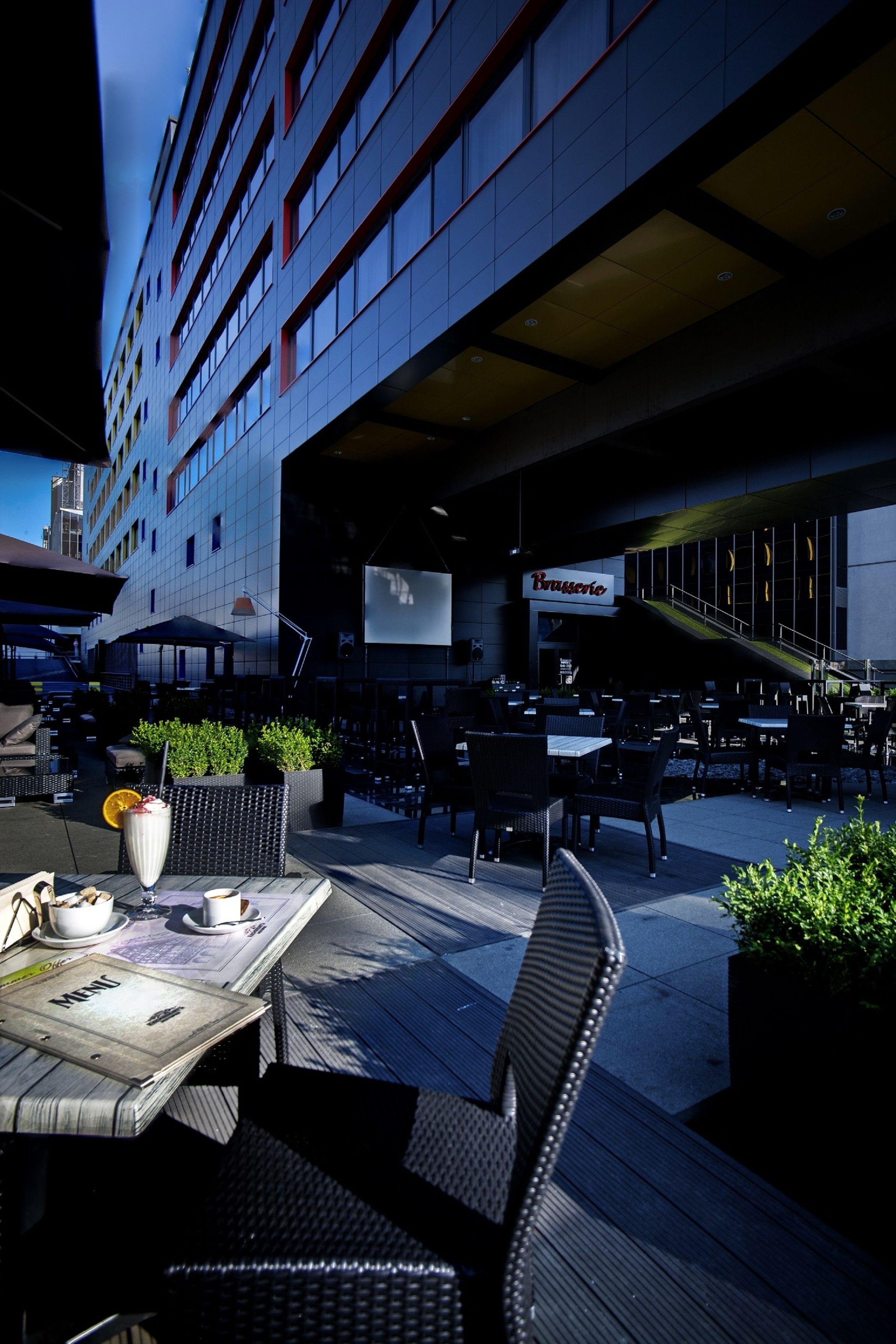 An outdoor seating area with tables and chairs, a sign for 'Brasserie', and a staircase leading to an upper level.