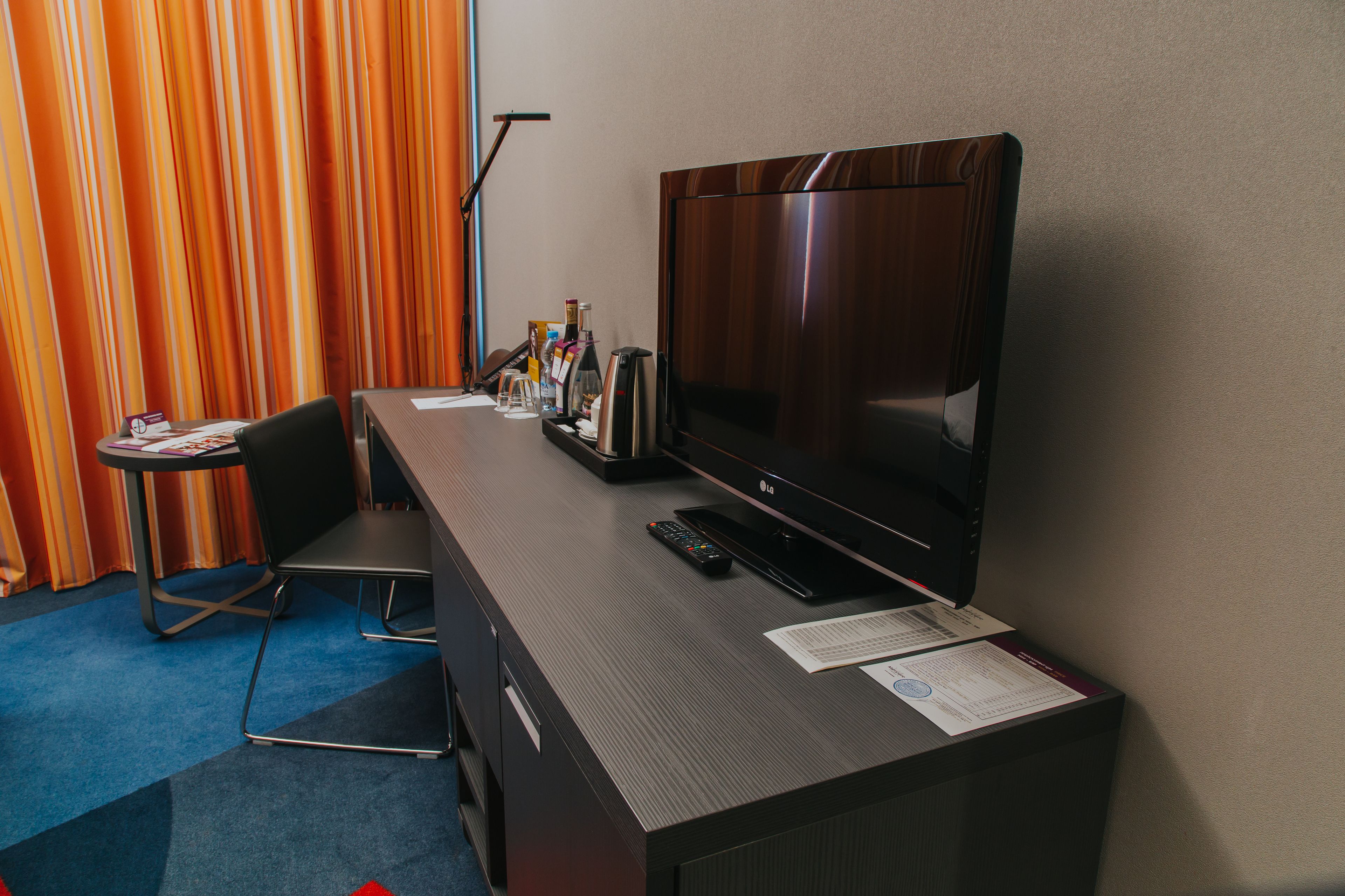 Hotelkamer met flatscreen-televisie, bureau, stoel, klein tafeltje en gordijn.