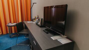 Hotelkamer met flatscreen-televisie, bureau, stoel, klein tafeltje en gordijn.