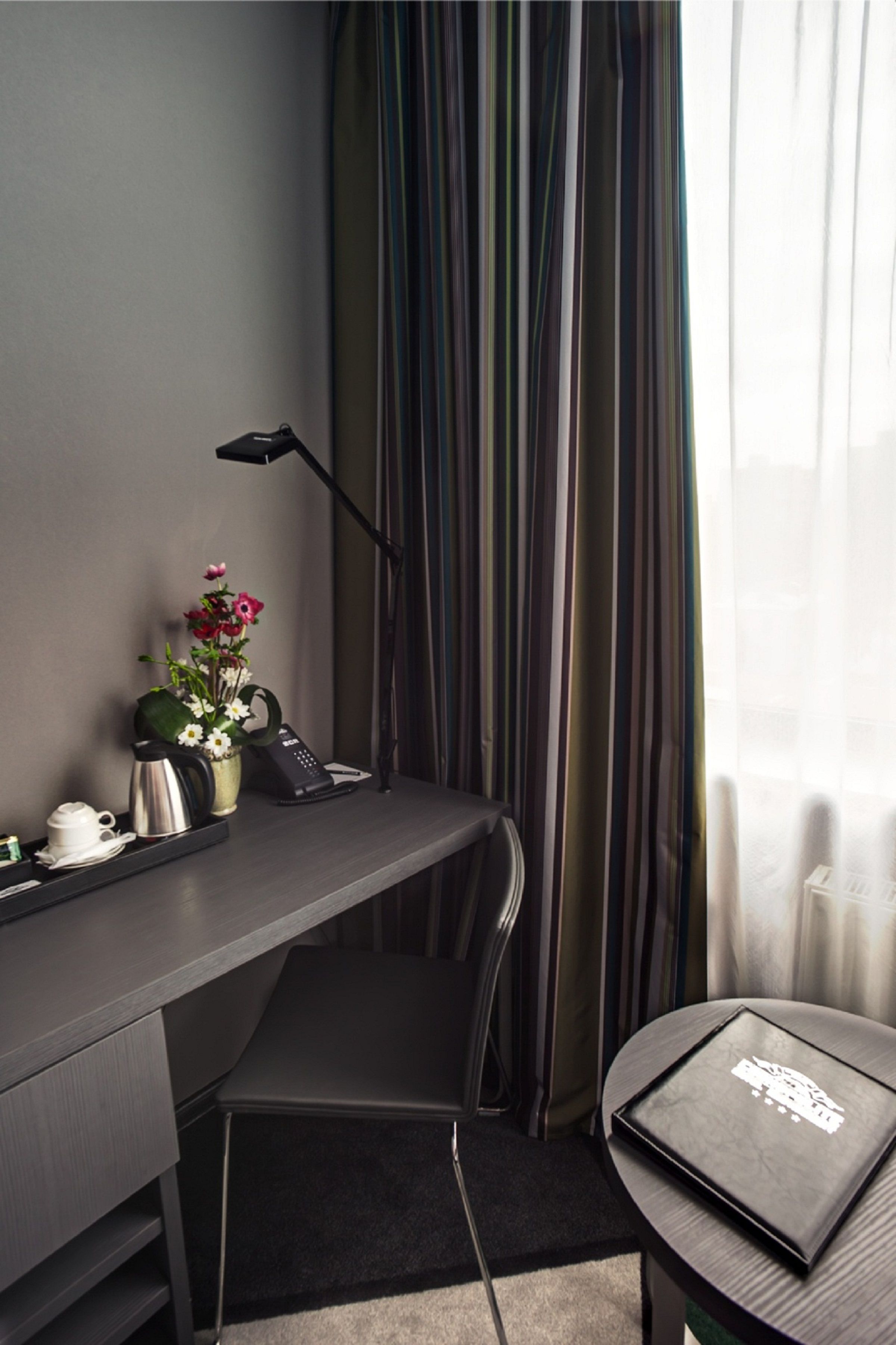 A modern hotel room with a desk, a chair, a coffee maker, a vase with flowers, a telephone, and a laptop on a small table.