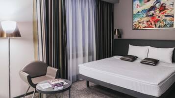 A hotel room with a bed, a chair, a small table, a lamp, and a painting on the wall.
