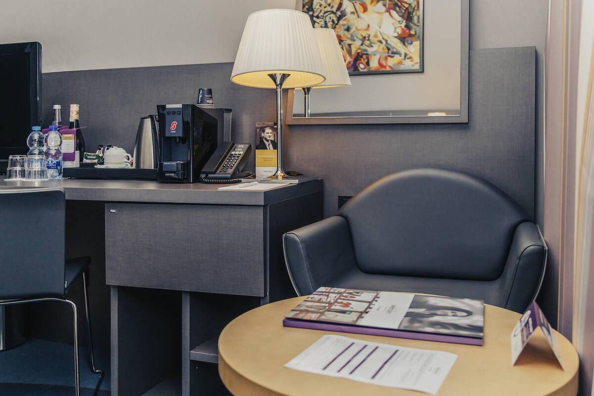 Ett hotellrum med ett skrivbord, en stol, en lampa, en kaffemaskin och ett litet bord med en tidning och några papper.