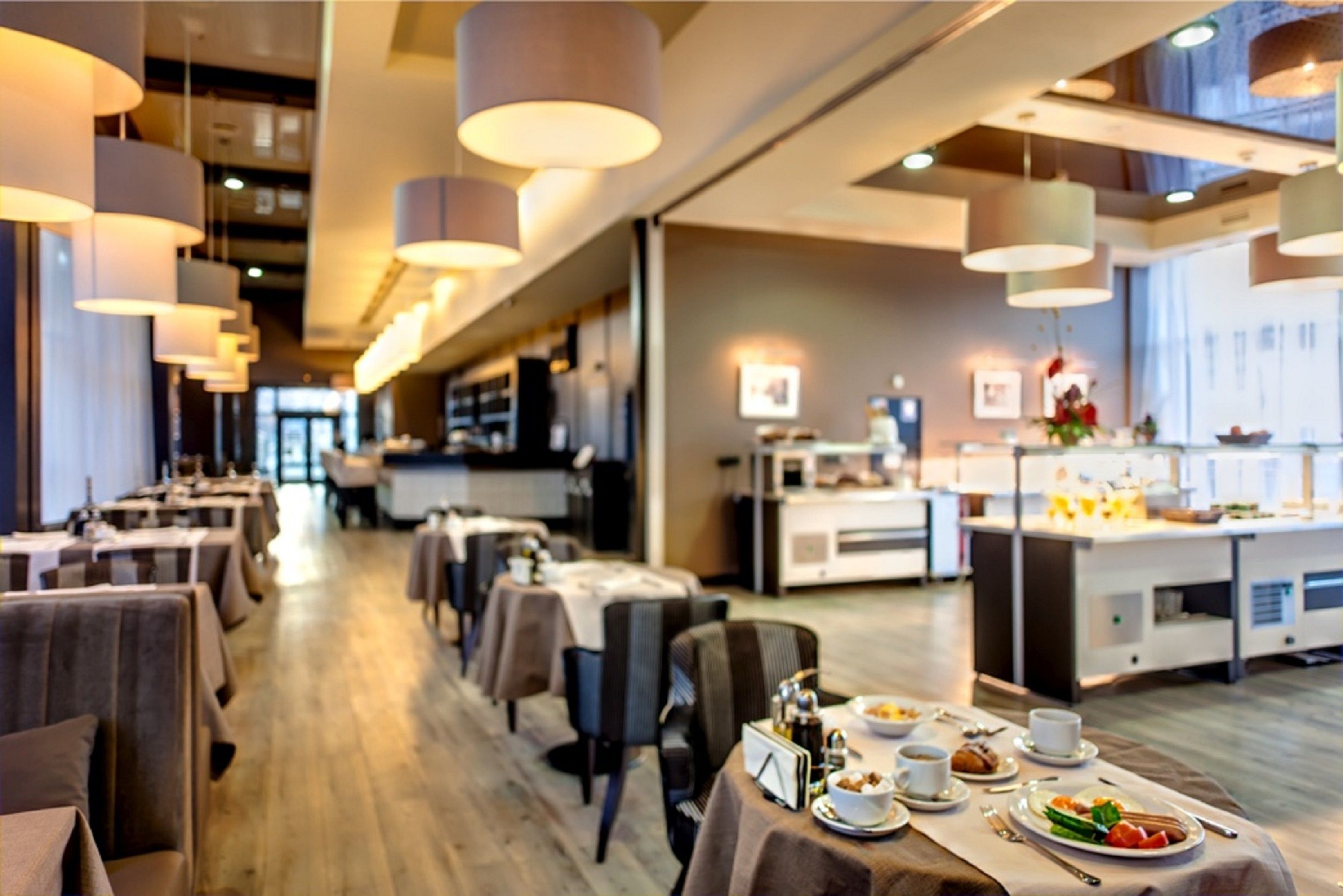 A modern restaurant with long tables, hanging pendant lights, and a kitchen area visible in the background.