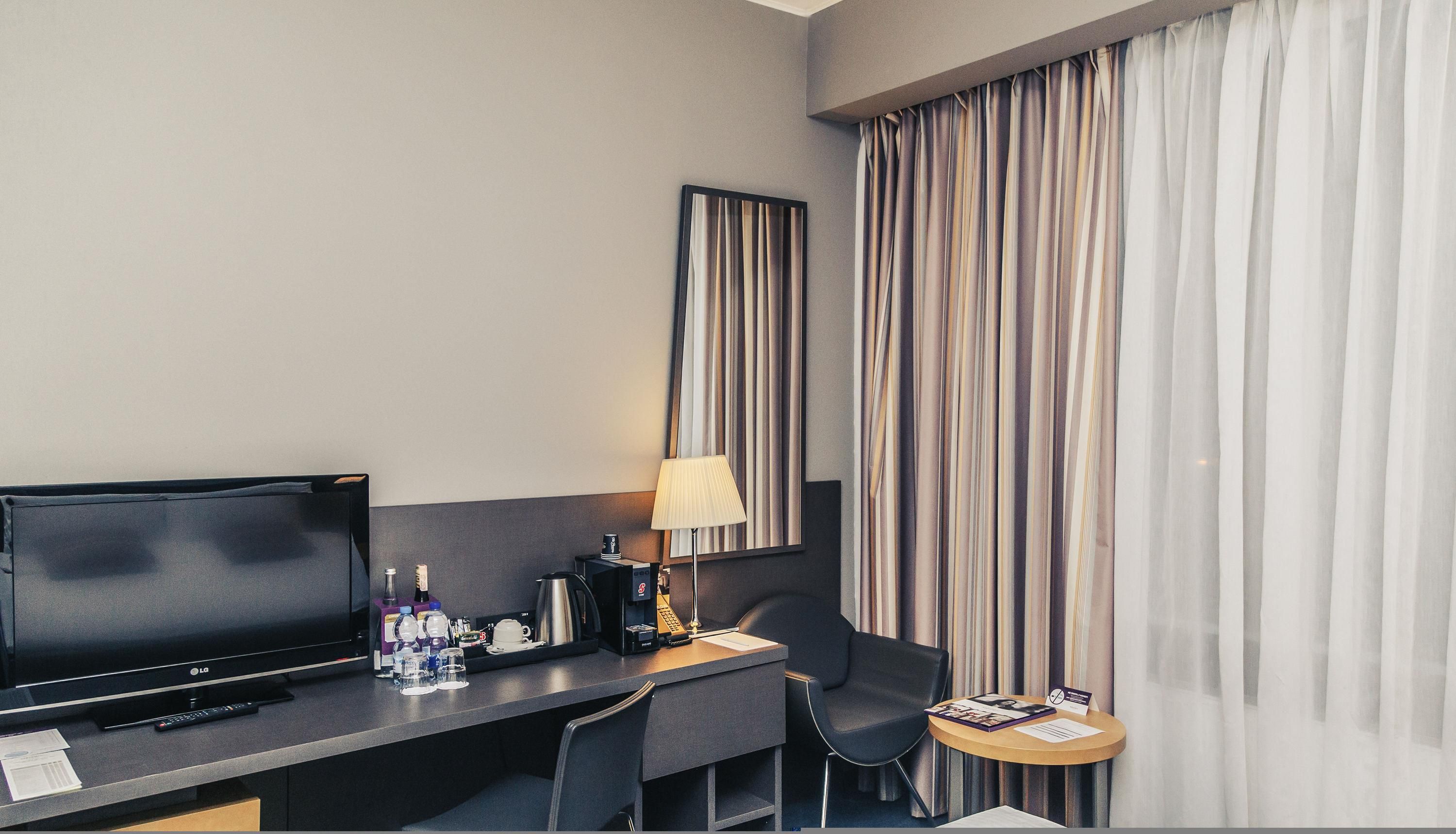 A hotel room with a flat-screen TV, a desk with a lamp, a chair, a small table with a brochure, and a large window with striped curtains.