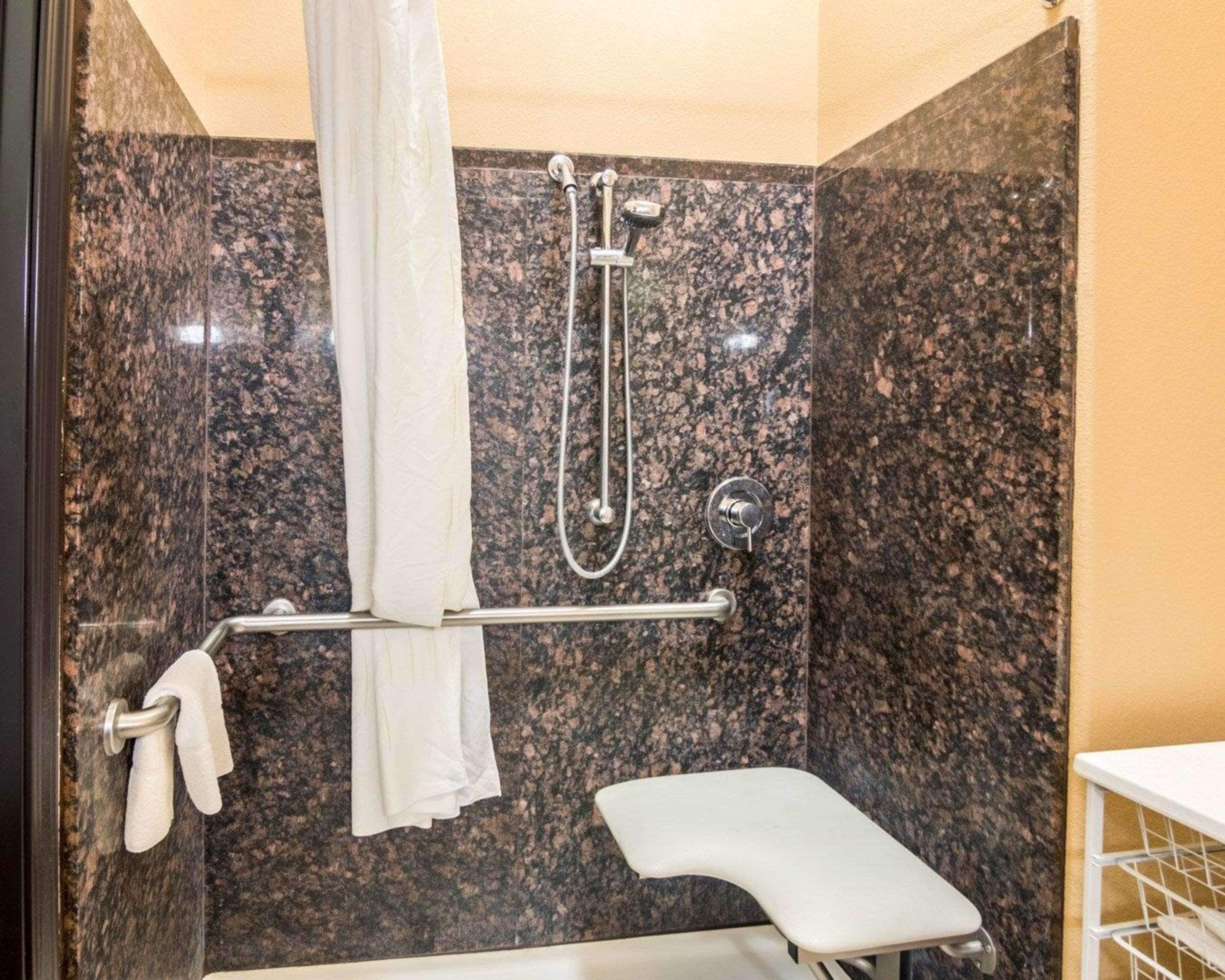 A bathroom with a granite shower wall, a showerhead, a towel rack with towels, and a white bench.