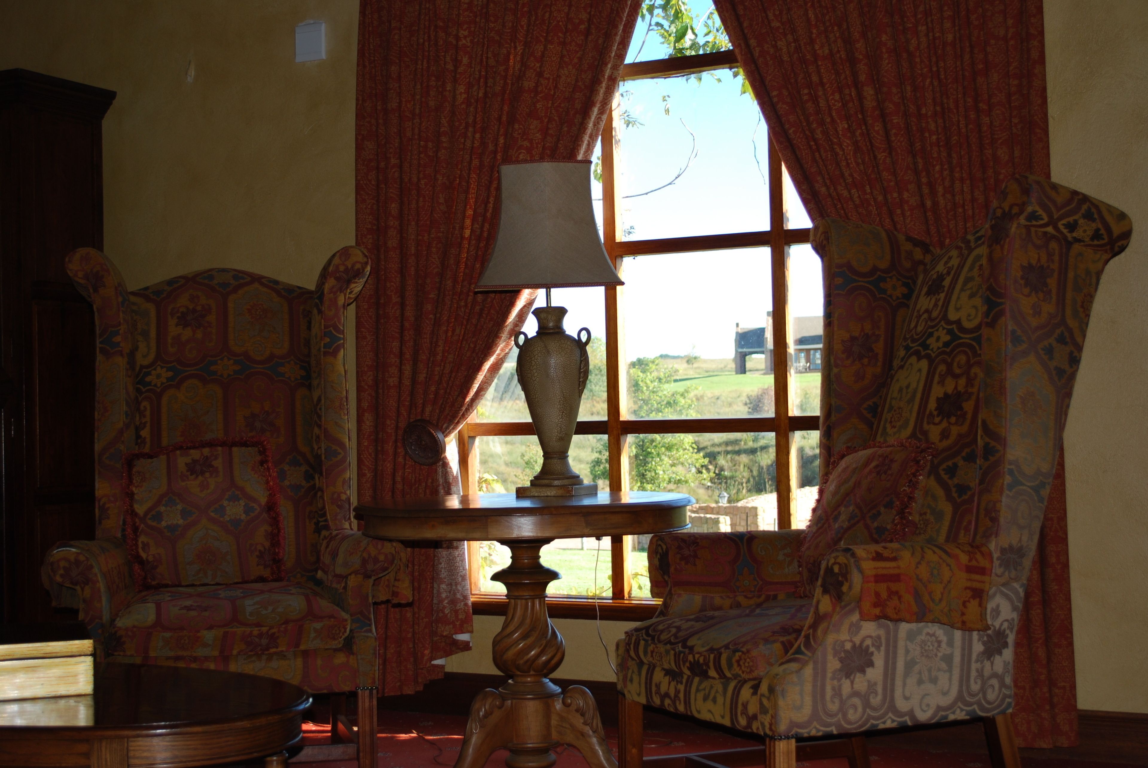 A room with two patterned armchairs, a wooden table with a lamp, and a window with curtains.