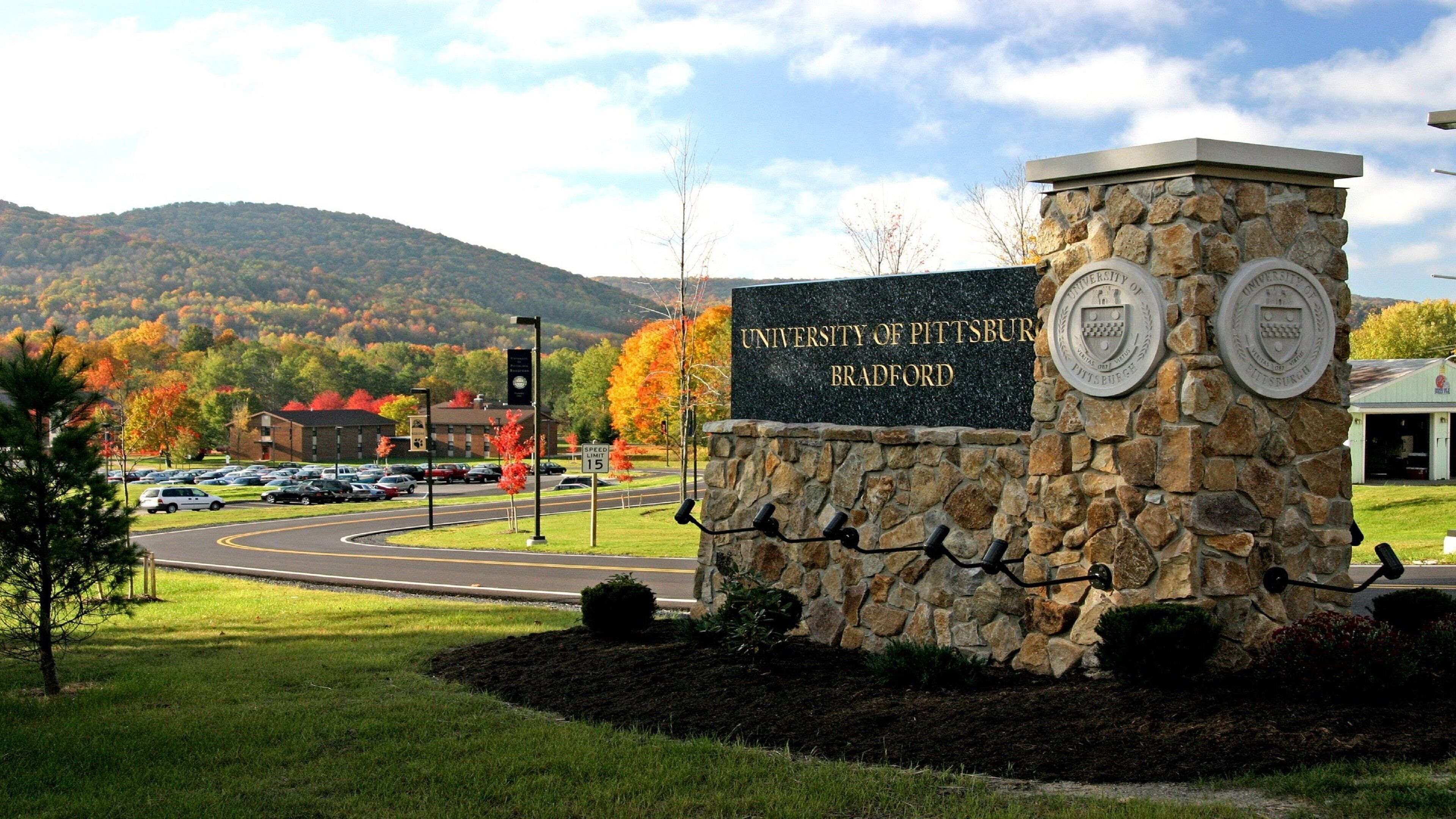A stone sign with the University of Pittsburgh Bradford name, surrounded by a landscaped area with grass and trees, a road with a speed limit sign, and a mountain in the background.