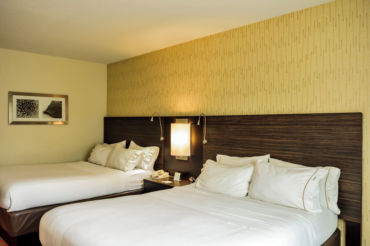 A hotel room with two beds, a nightstand, a wall-mounted lamp, and a framed picture.