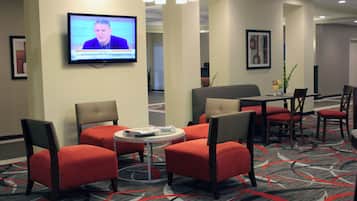 Un hall de hotel con un televisor mostrando un noticiero, asientos tapizados en rojo, una pequeña mesa redonda con una revista y una alfombra estampada.