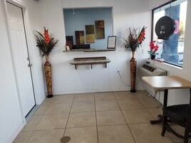 Un área de recepción con suelo de baldosas, mostrador de recepción, dos jarrones con flores, obras de arte enmarcadas en la pared y una ventana con vistas al exterior.