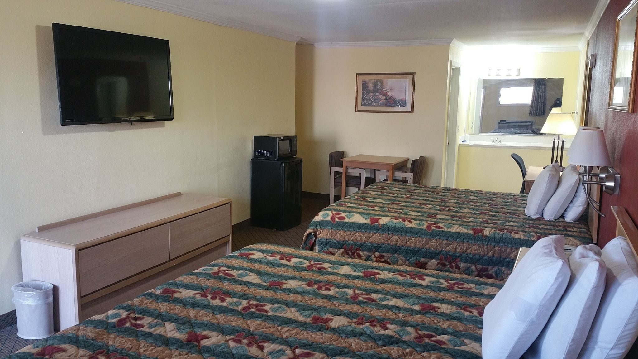 Una camera d'albergo con due letti, una televisione, una scrivania, una sedia, un cestino e un quadro appeso al muro.