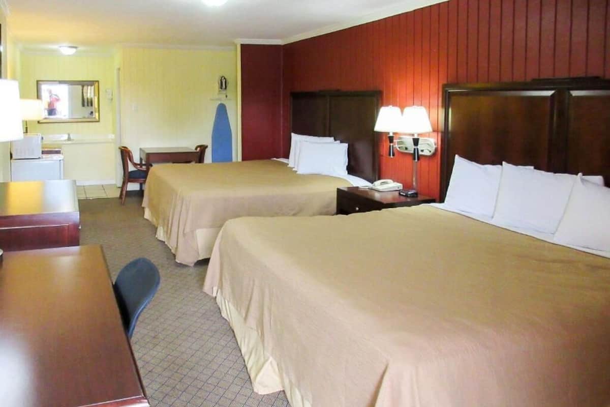 A hotel room with two beds, a desk, a chair, a small table, a mirror, and a wall-mounted lamp.