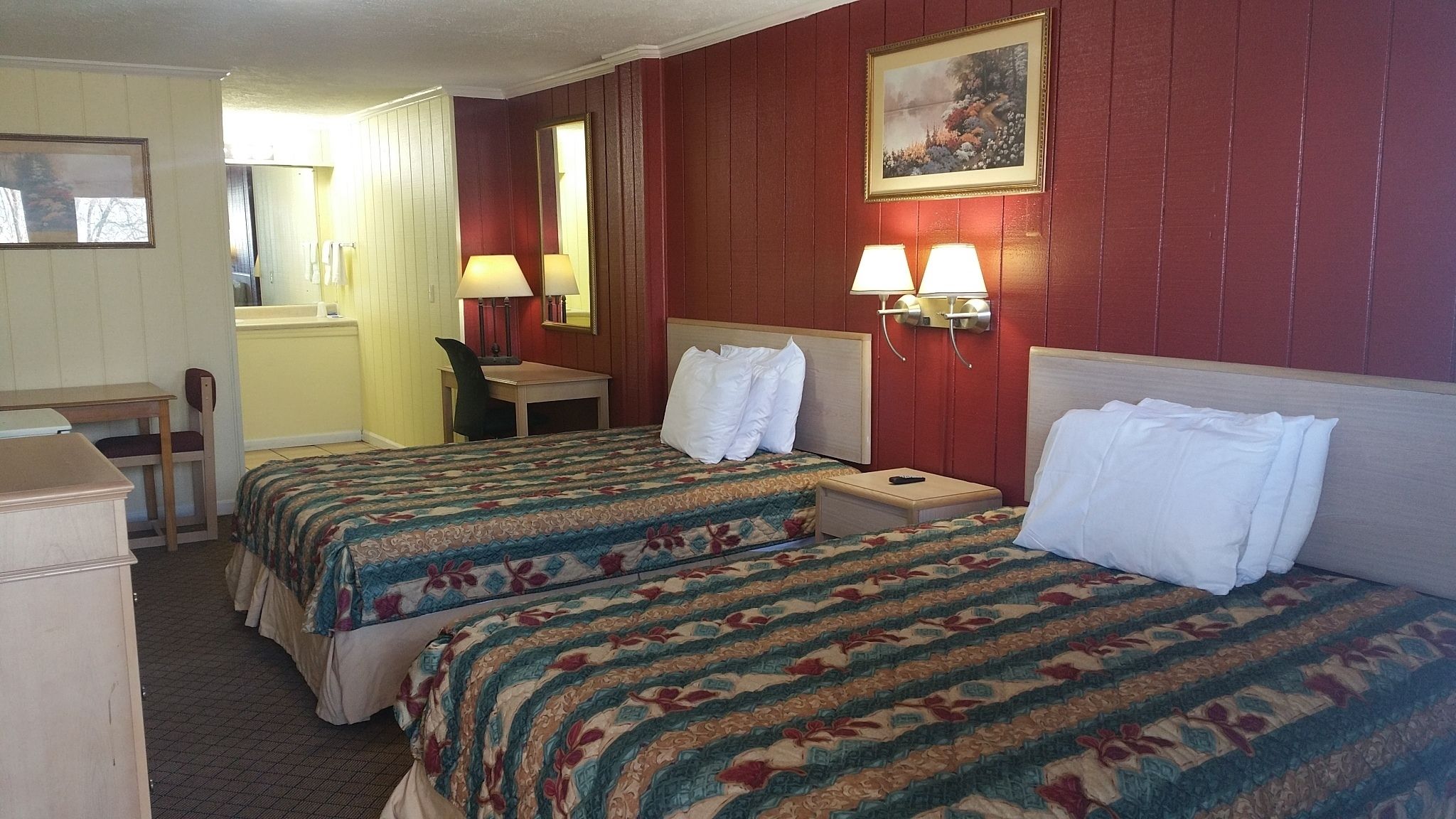 Quarto de hotel com duas camas, uma escrivaninha, uma cadeira, um abajur e um quadro na parede.