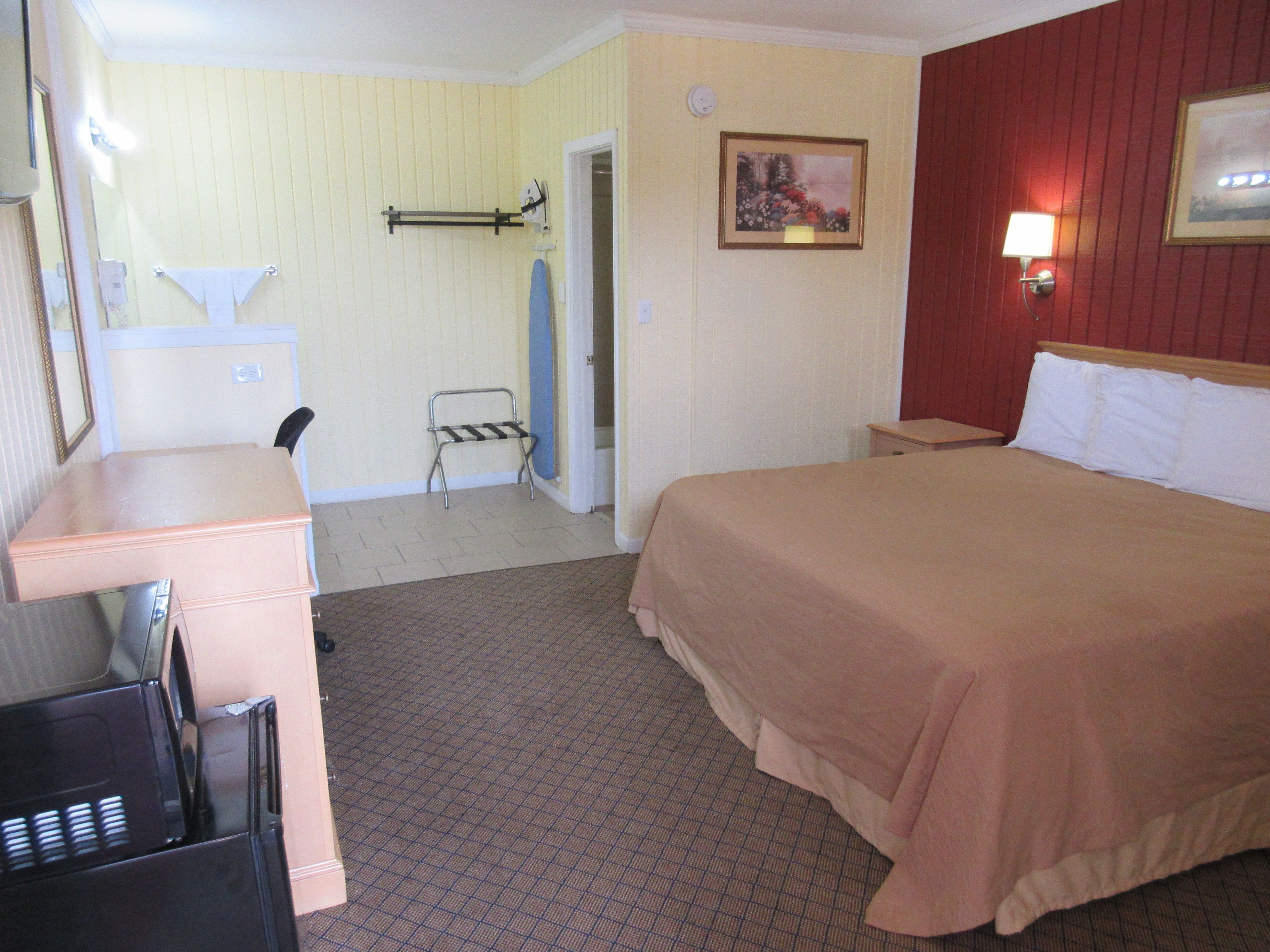 A hotel room with a bed, a desk, a chair, a coat rack, and a bathroom visible through an open door.
