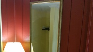 Ein kleines Zimmer mit Schreibtisch, Stuhl, Lampe, Spiegel und Mülleimer.