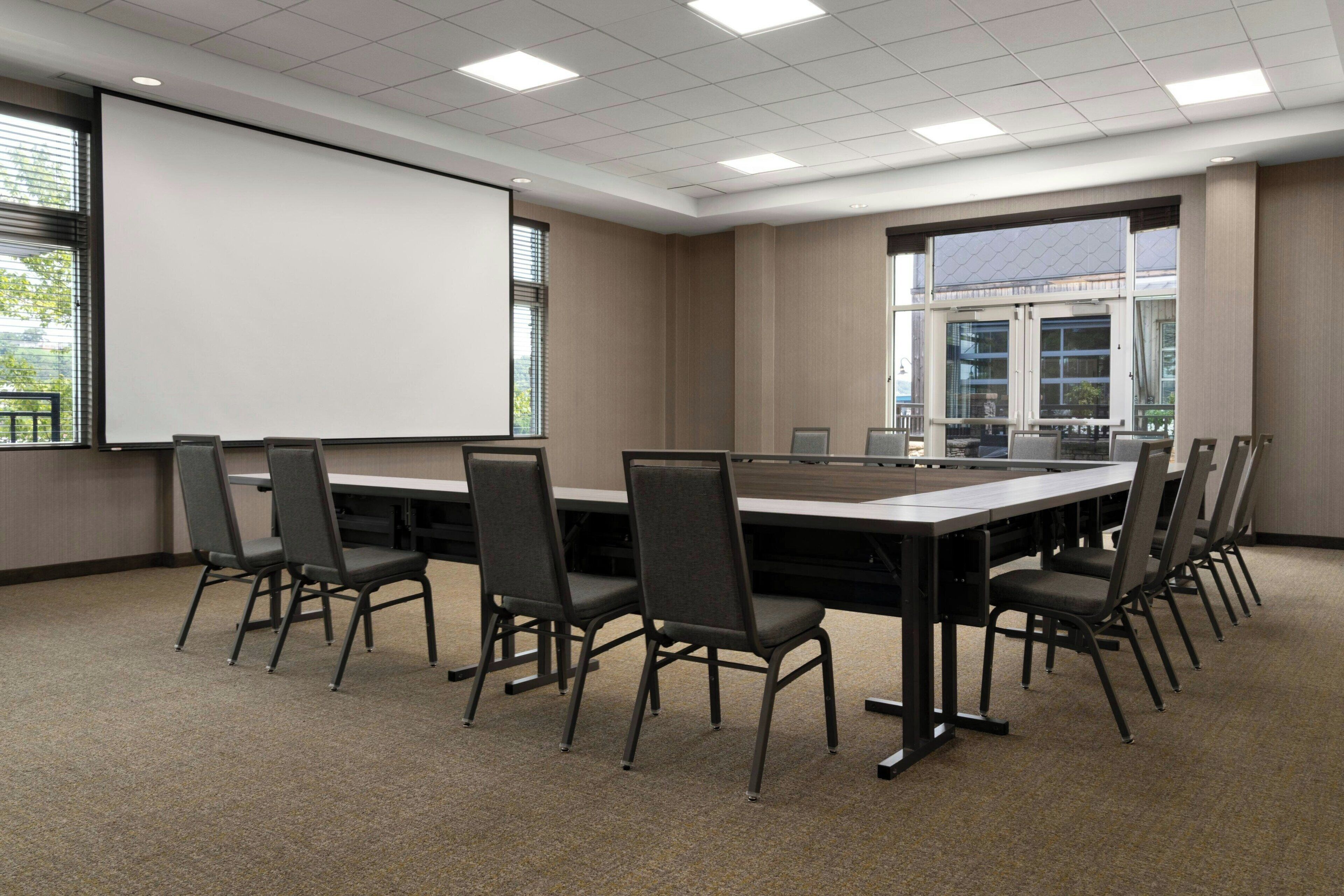 Un salón de conferencias con una larga mesa, sillas, una pantalla de proyector y ventanas amplias.