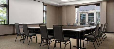 Un salón de conferencias con una larga mesa, sillas, una pantalla de proyector y ventanas amplias.