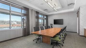 A conference room with a large table, chairs, a flat-screen TV, and large windows offering a view of buildings and a body of water.