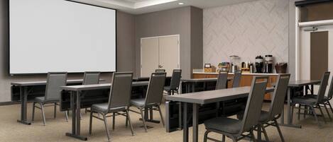 A conference room with tables and chairs, a projector screen, a coffee station, and a door.