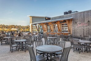An outdoor patio with multiple round tables and chairs, a wooden building with a neon sign, and a view of a hilly landscape in the background. - SpringHill Suites by Marriott Chattanooga Downtown/Cameron Harbor (Chattanooga)