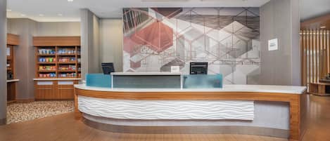 Un espace d’accueil d’hôtel au design moderne, comprenant un comptoir de réception, un écran numérique fixé au mur et un bar proposant divers produits emballés.