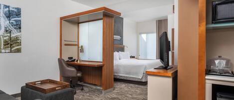 A hotel room with a bed, a desk with a chair, a television, a small kitchenette, and a painting on the wall.