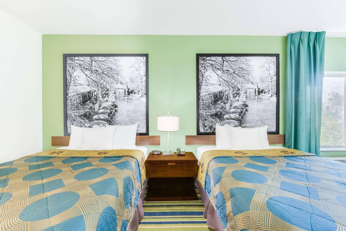 A hotel room with two beds, a nightstand, a lamp, and two large black-and-white photographs on the wall.