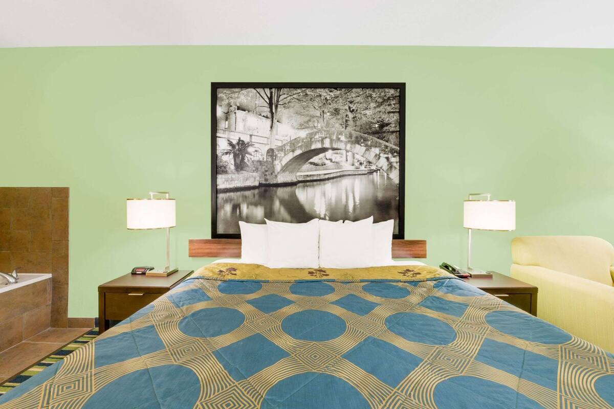 A hotel room with a bed, two bedside lamps, a chair, a nightstand, a bathroom with a bathtub, and a large framed picture on the wall.