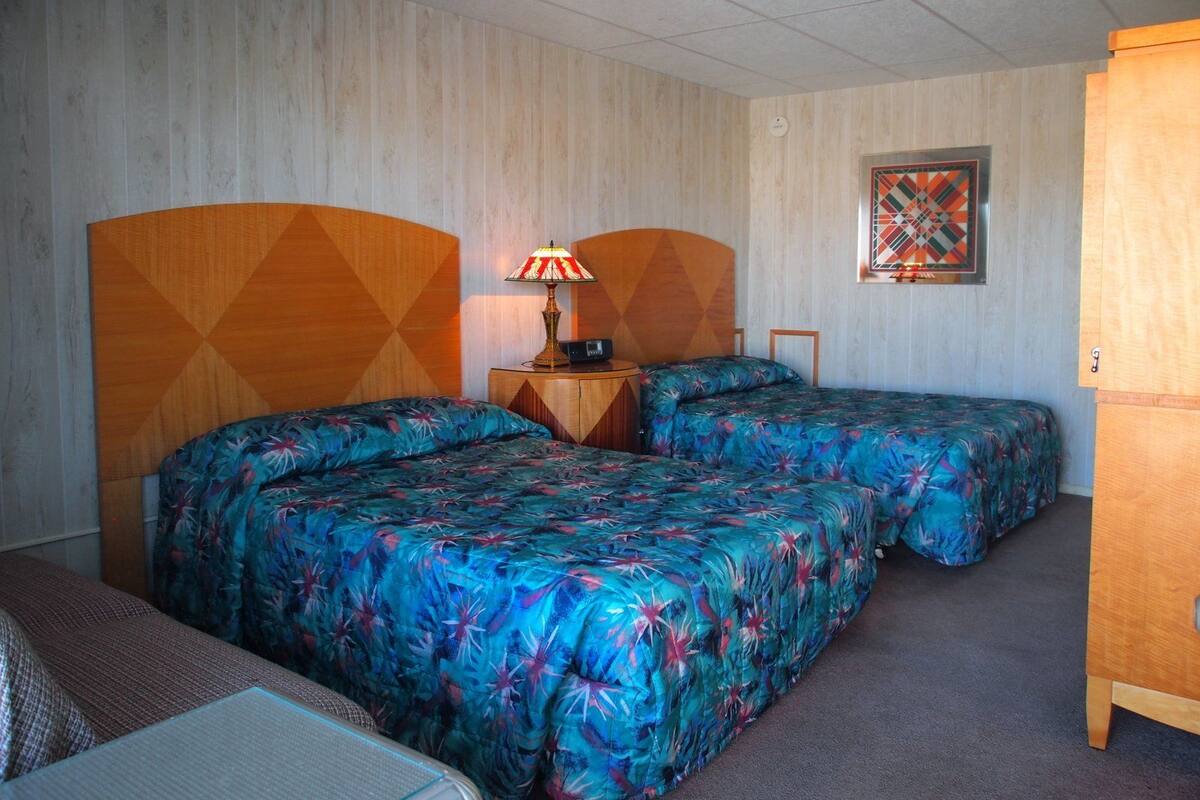 A hotel room with two beds, a nightstand, a lamp, a clock, and a painting on the wall.