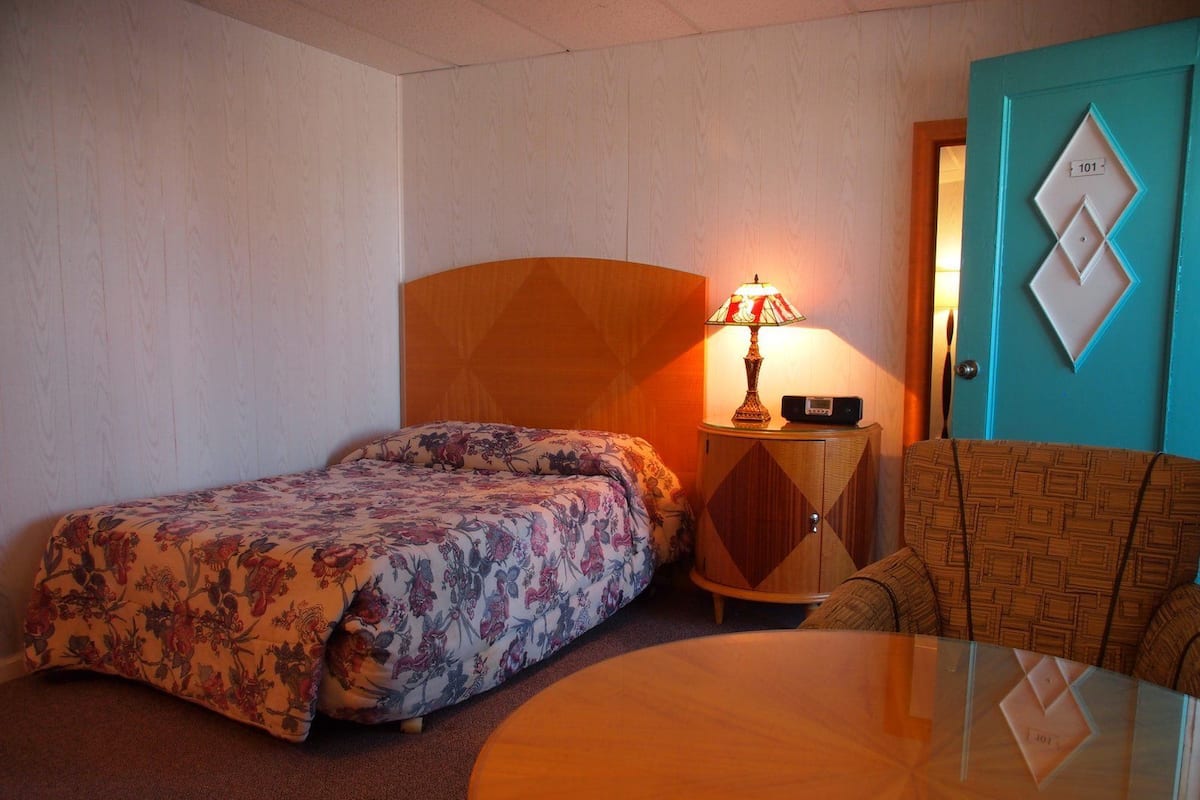 A hotel room with a bed, a round table, a chair, a lamp, and a door with a number.