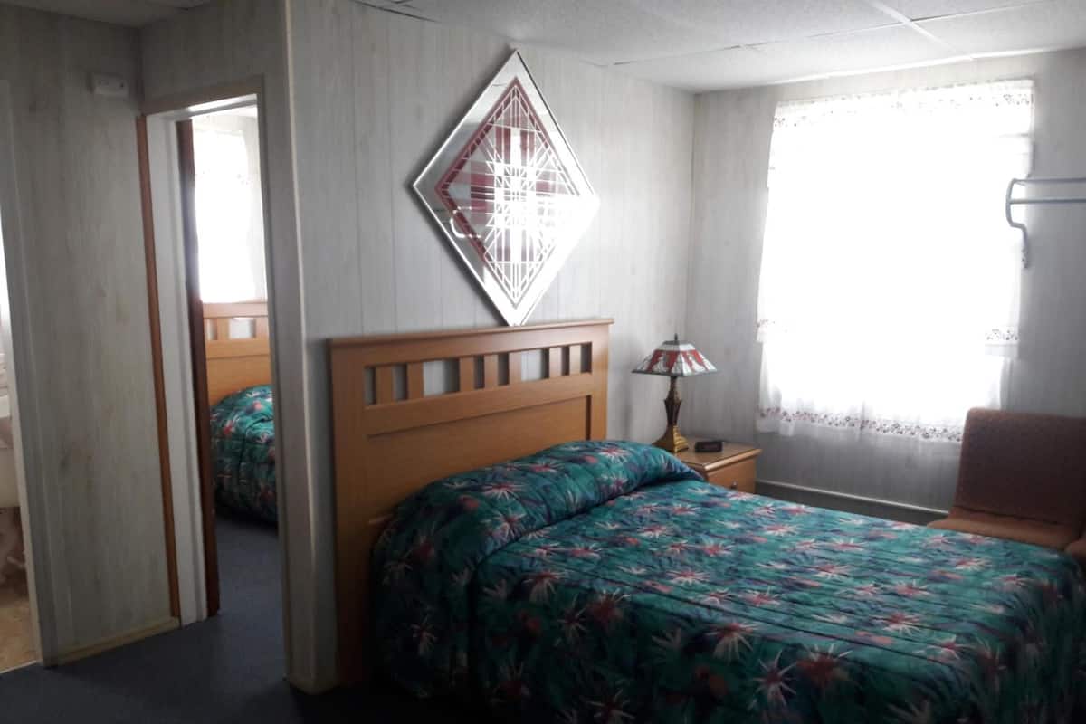 A bedroom with a wooden bed, a nightstand with a lamp, a window with sheer curtains, and a mirror on the wall.