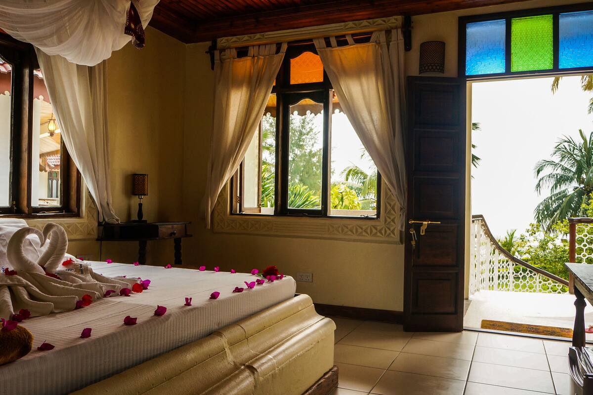 A hotel room with a bed adorned with rose petals, a decorative towel, and a swan-shaped pillow. There is a window with curtains, a nightstand with a lamp, and a view of greenery outside.