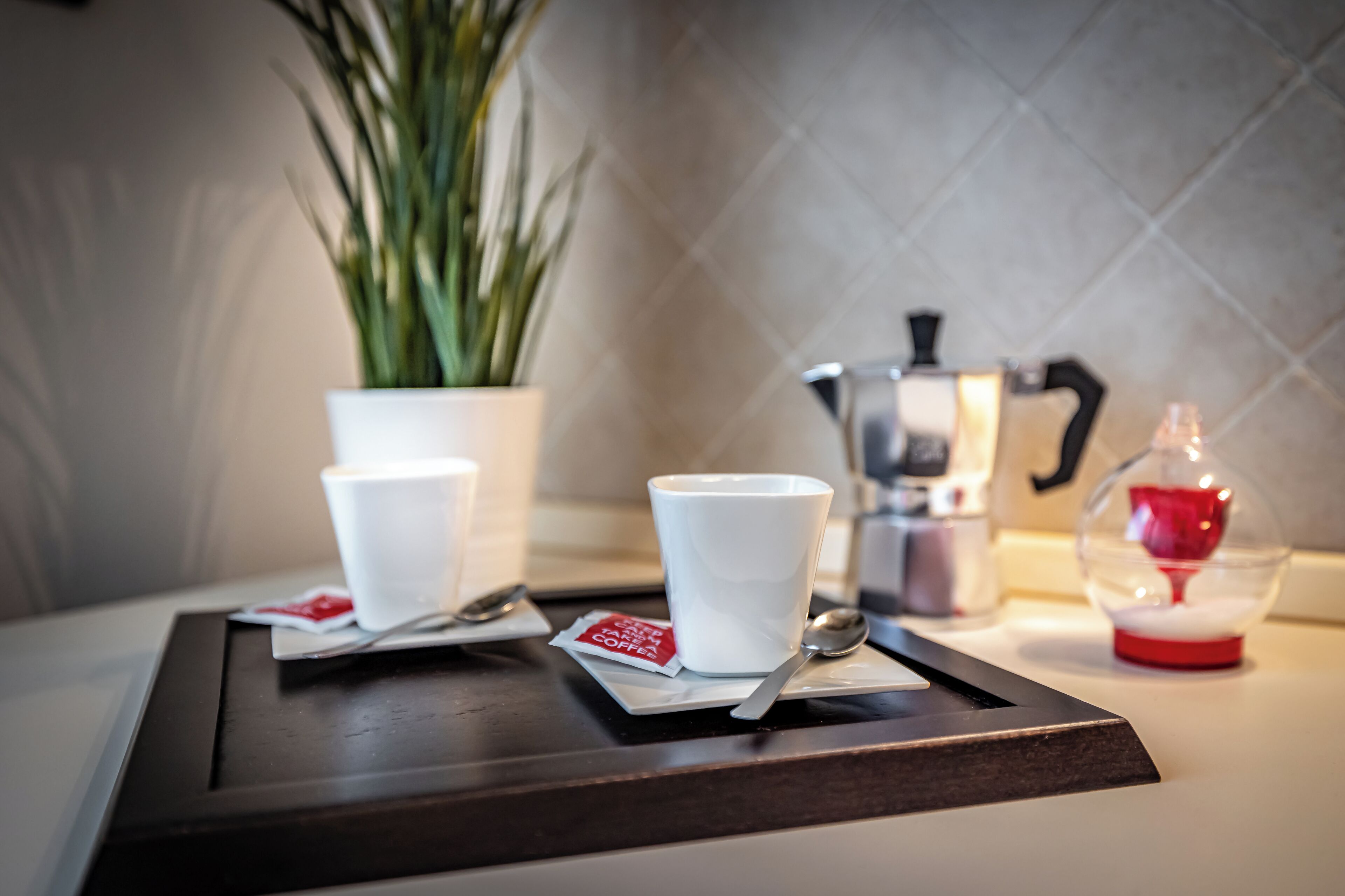 Ein Tablett mit zwei weißen Tassen, einem Löffel, einer Kaffeepackung, einer Pflanze, einer Kaffeemaschine und einem dekorativen Gegenstand auf einem Tisch.