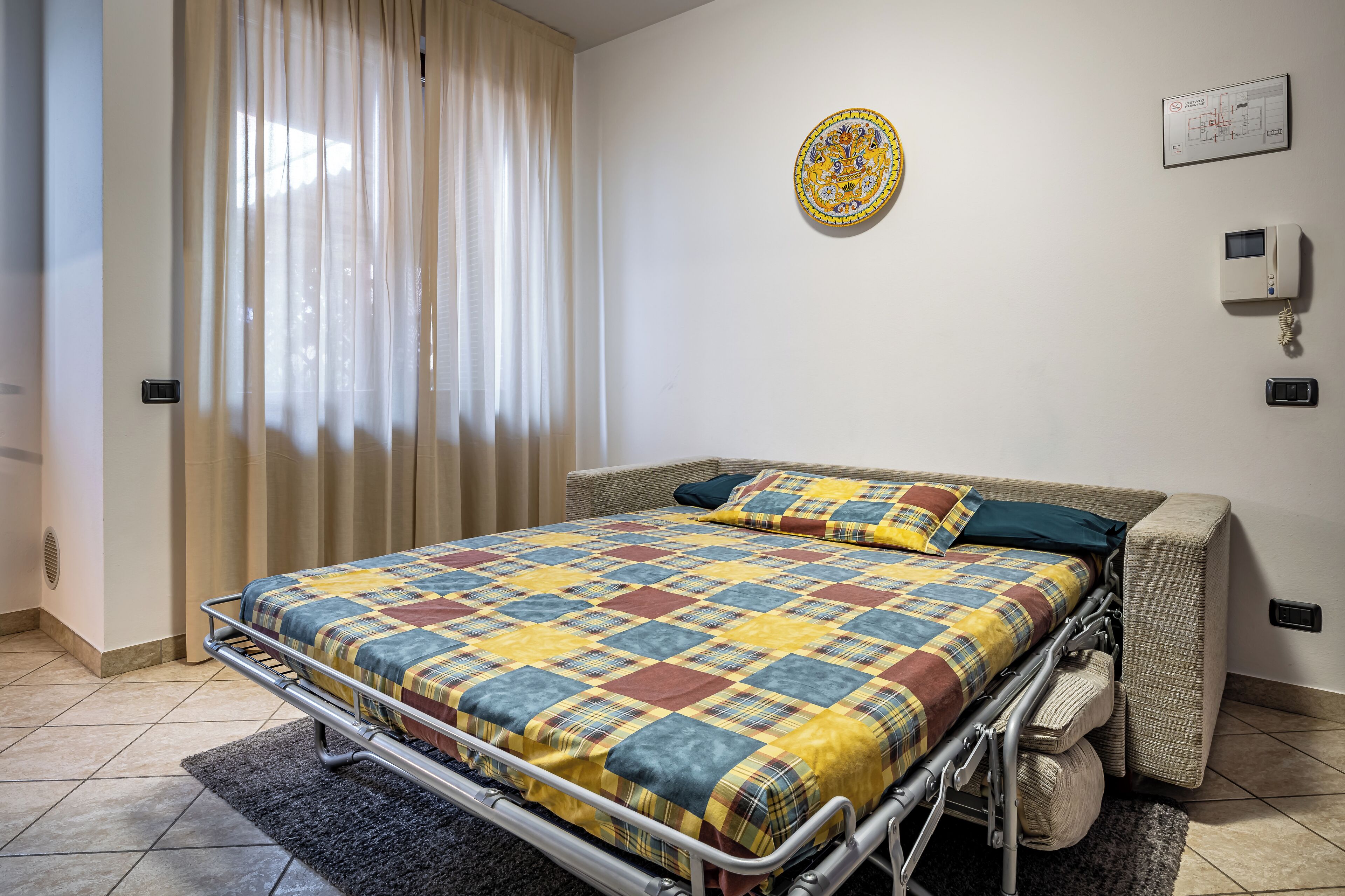 Un dormitorio con estructura de cama metálica, colcha acolchada, ventana con cortinas de gasa, termostato en la pared y una alfombra pequeña en el suelo.