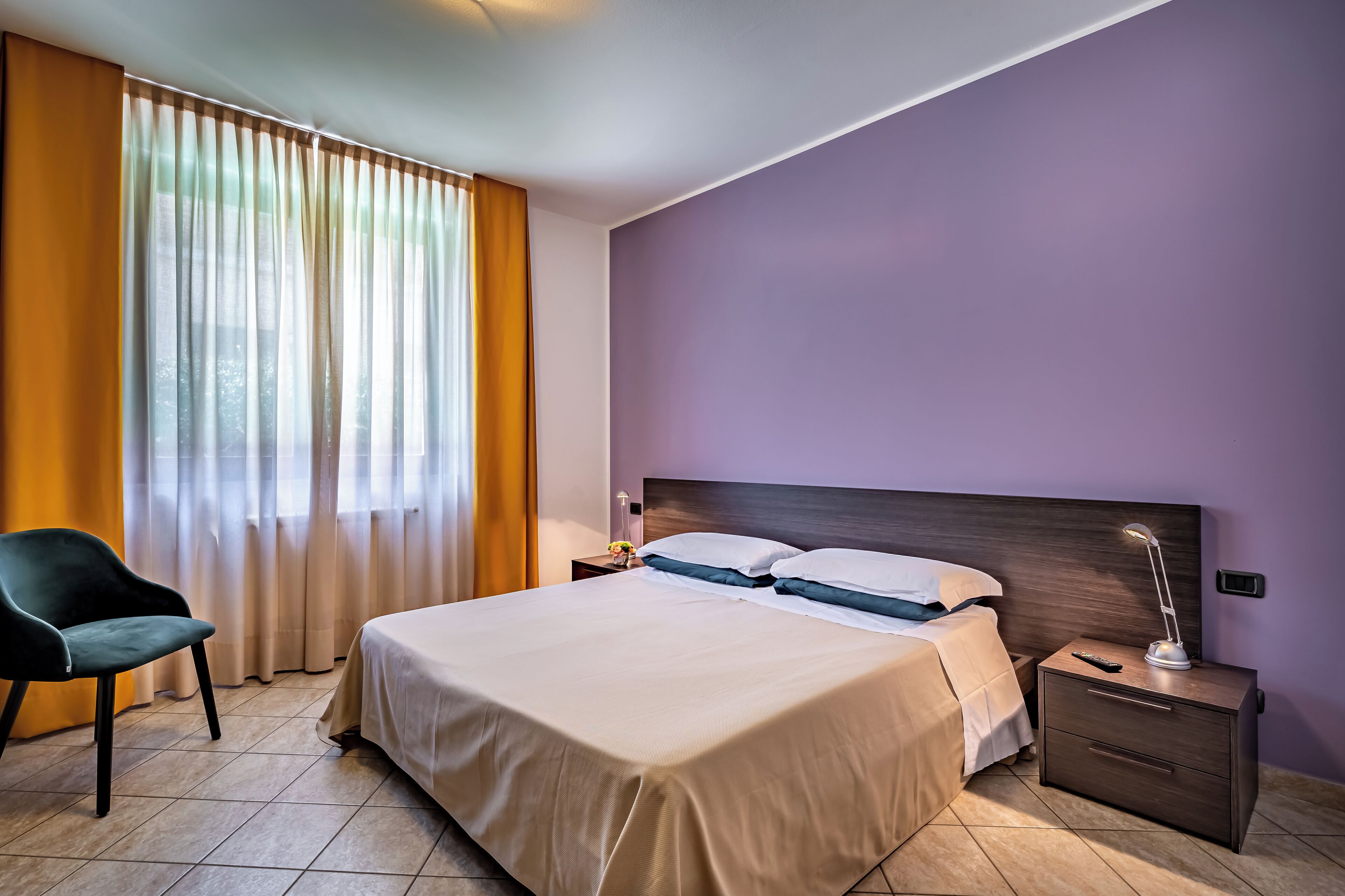 A hotel room with a bed, nightstand, lamp, chair, and curtains.