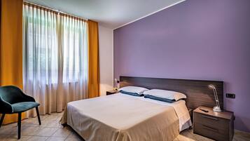 A hotel room with a bed, nightstand, lamp, chair, and curtains.