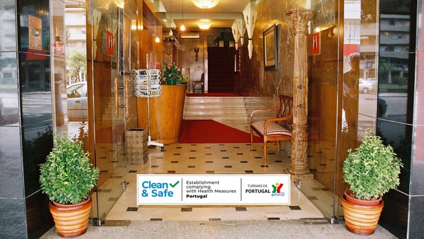 Entrada de hotel con mostrador de recepción, alfombra roja y dos plantas en macetas. Un cartel indica el cumplimiento de medidas sanitarias.