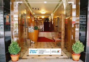 A hotel entrance with a reception desk, a red carpet, and two potted plants. A sign indicates compliance with health measures. - Hotel Vice-Rei (Porto)