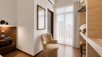 Quarto de hotel com piso de madeira, uma poltrona bege, uma pequena mesa de trabalho com telefone, um espelho e uma janela com cortinas translúcidas.