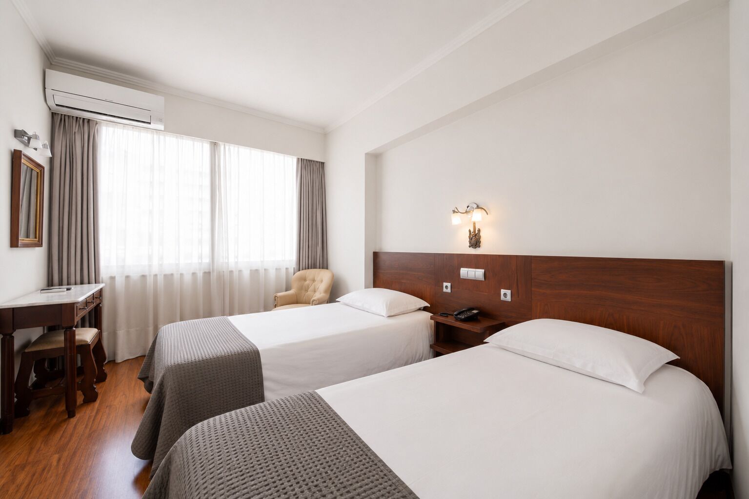 Quarto de hotel com duas camas, uma escrivaninha, uma cadeira e mesa de cabeceira com telefone.