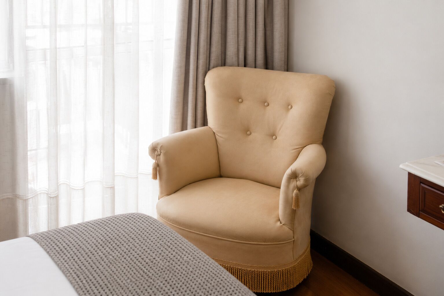 A beige armchair with tufted backrest and armrests, positioned next to a bed with a white headboard and a grey knitted throw.