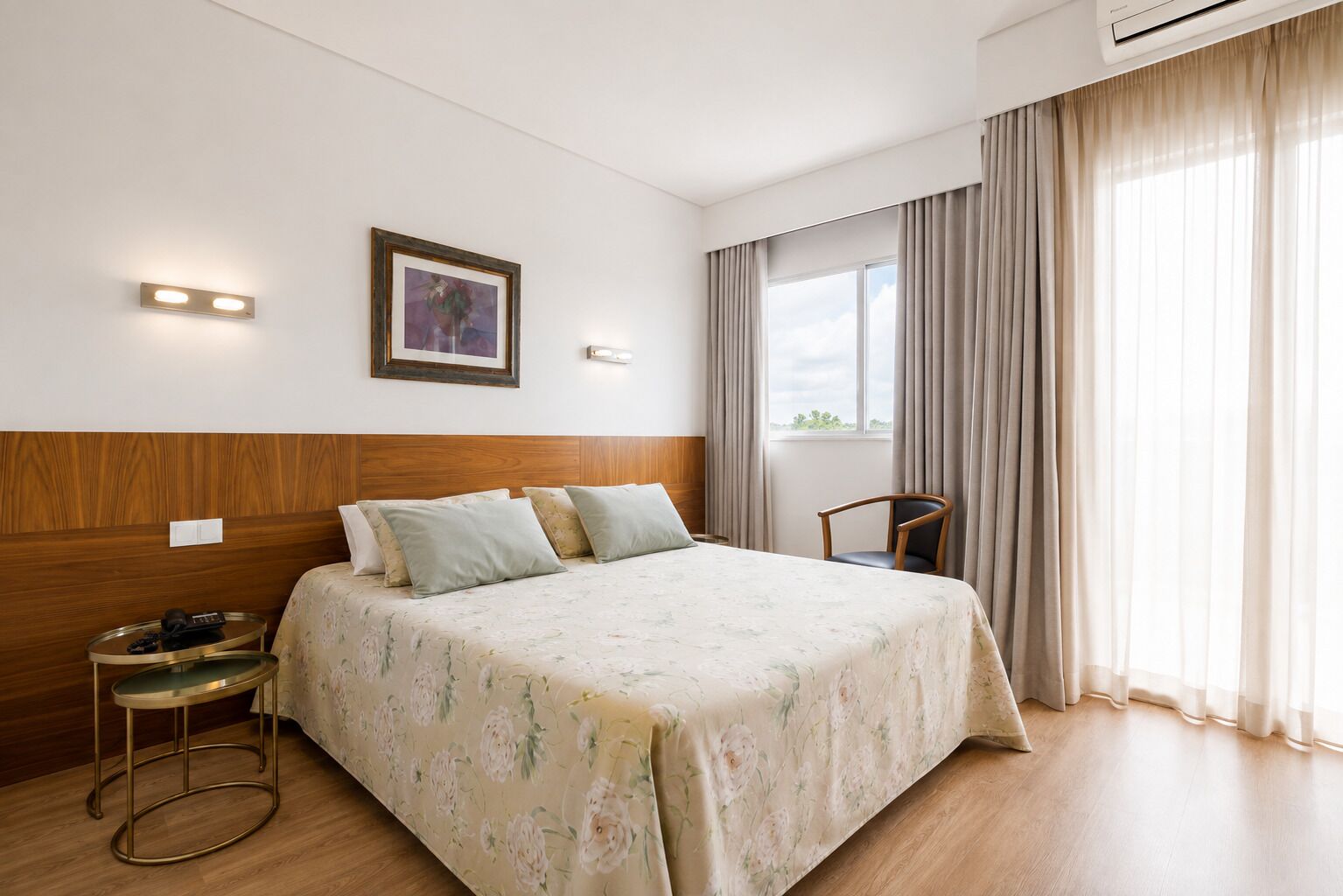 A hotel room with a wooden headboard, a bed with floral bedding, a round side table, a chair, a window with curtains, and a wall-mounted air conditioning unit.