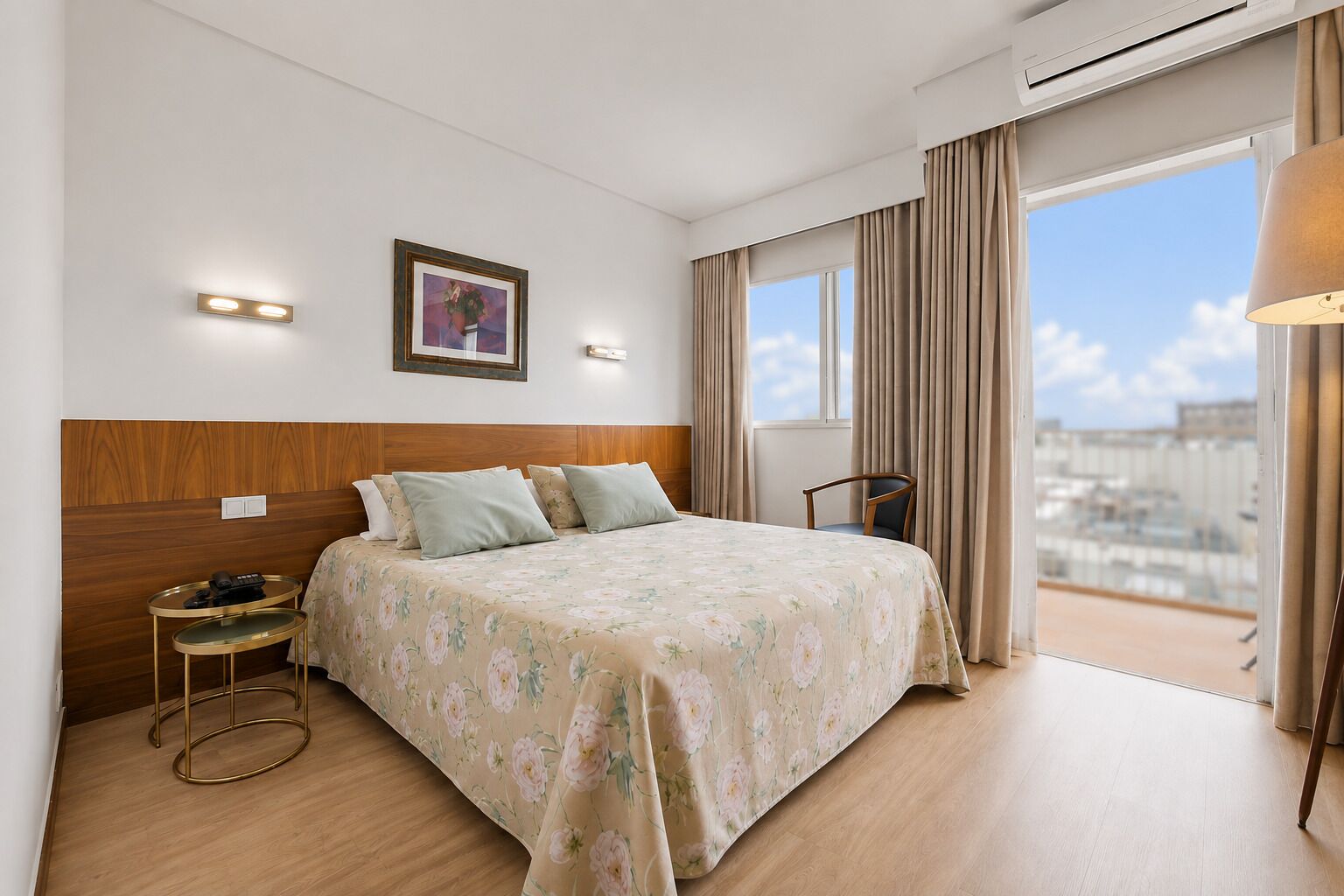 A hotel room with a bed, bedside table, lamp, and a view of the city.