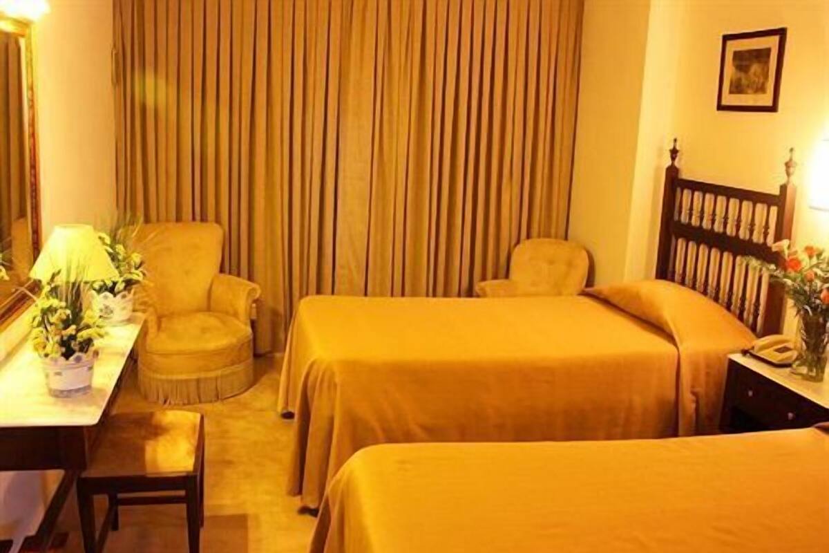 Une chambre d’hôtel avec deux lits simples, un bureau avec une lampe, une chaise et une grande fenêtre avec des rideaux.