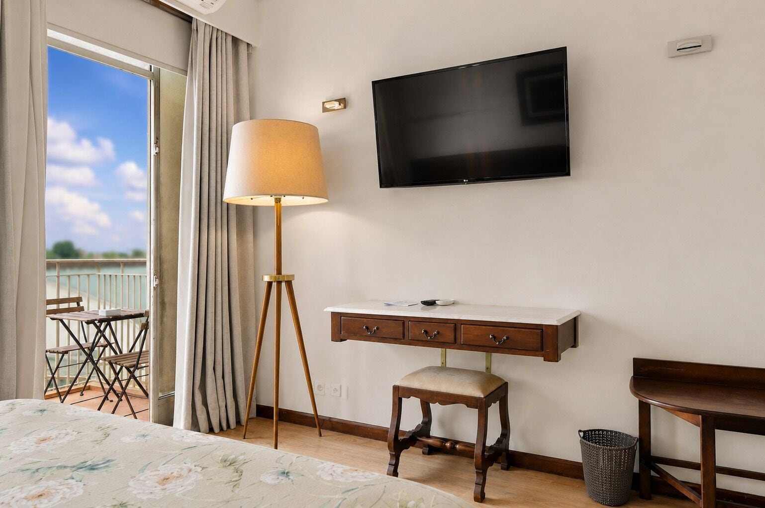 Habitación de hotel con cama, escritorio, silla, lámpara de pie, televisión y balcón con sillas.