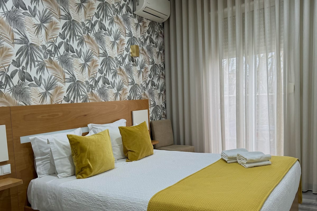Una cama bien tendida con ropa de cama blanca y amarilla, cabecera de madera y papel tapiz con estampado de hojas tropicales.