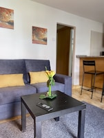 A living room with a dark square table, a sofa with yellow cushions, and a vase with flowers. There are two paintings on the wall and a door leading to another room.