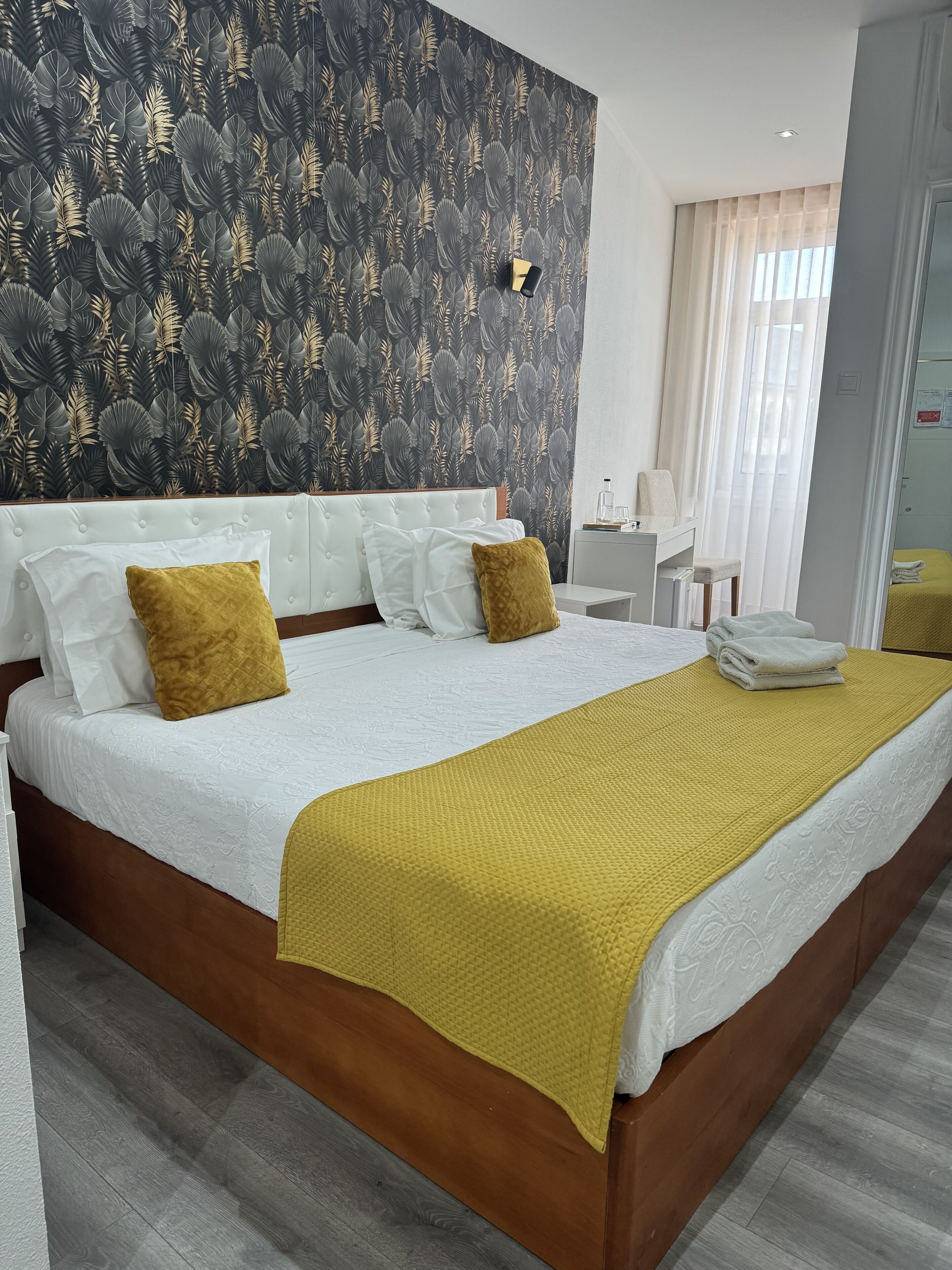 Ein Hotelzimmer mit einem großen Bett, einem Kopfteil, einem Nachttisch, einem Spiegel und einem Fenster mit Vorhängen.
