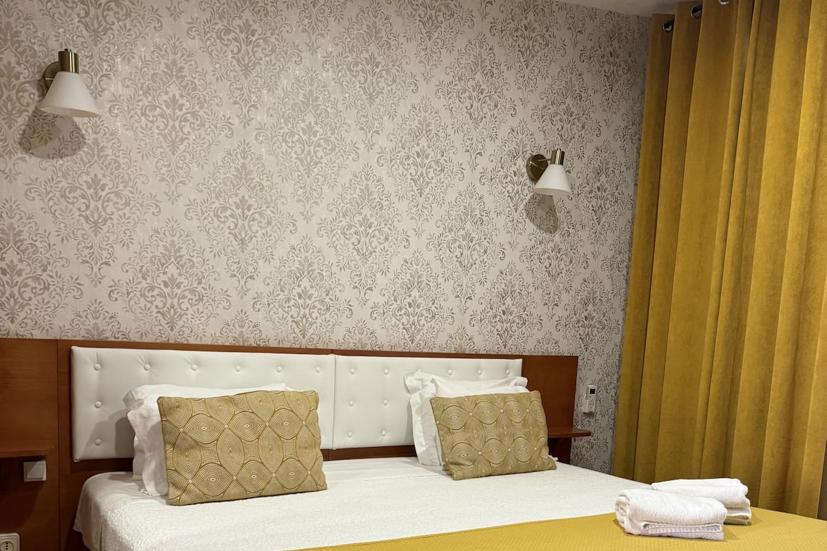 Una cama bien tendida con un edredón amarillo, dos almohadas estampadas y un cabecero blanco.